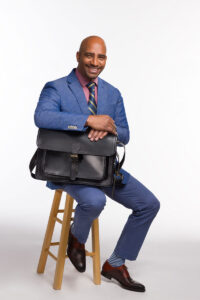 A smiling professional man in a blue suit, sitting on a stool and holding a black Brunswick leather briefcase