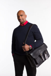 A professional man in black sweater and dress pants with a red shirt underneath, holding a black leather Brunswick briefcase