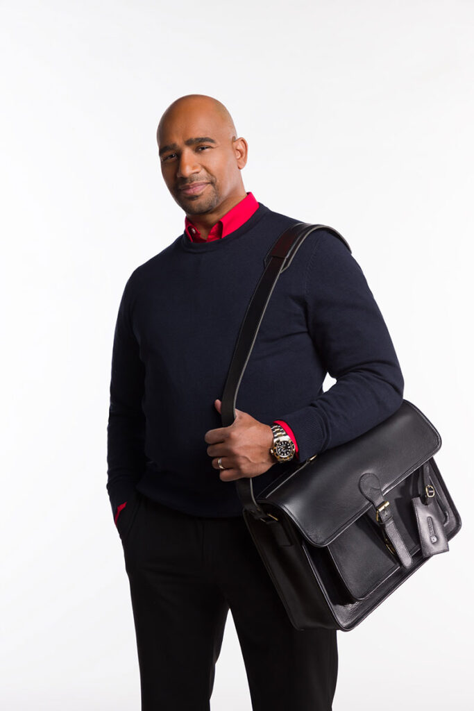 A professional man in black sweater and dress pants with a red shirt underneath, holding a black leather Brunswick briefcase