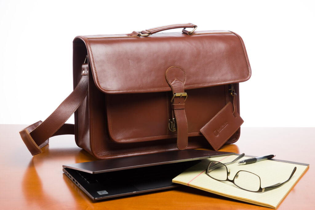 The Brunswick brown leather briefcase on a wooden desk, next to a legal pad, laptop, and a pair of glasses