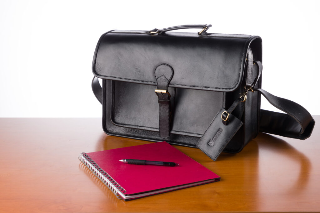 The Brunswick leather briefcase in black, on a wooden desk next to a red envelope and a black pen.